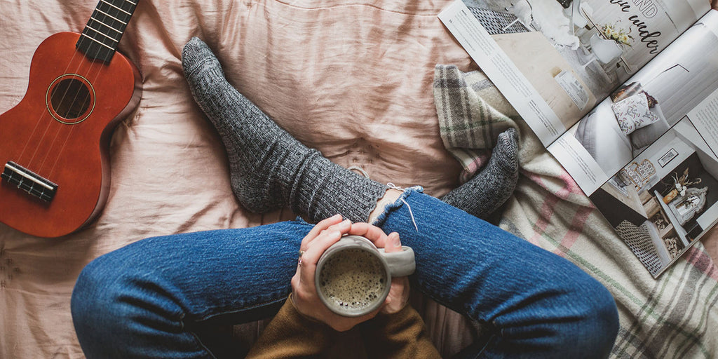Aerial top down view of person sitting cross legged wearing jeans & thick grey socks sitting on a duvet cover holding a mug of coffee, guitar on the left hand side and magazine on the right hand side